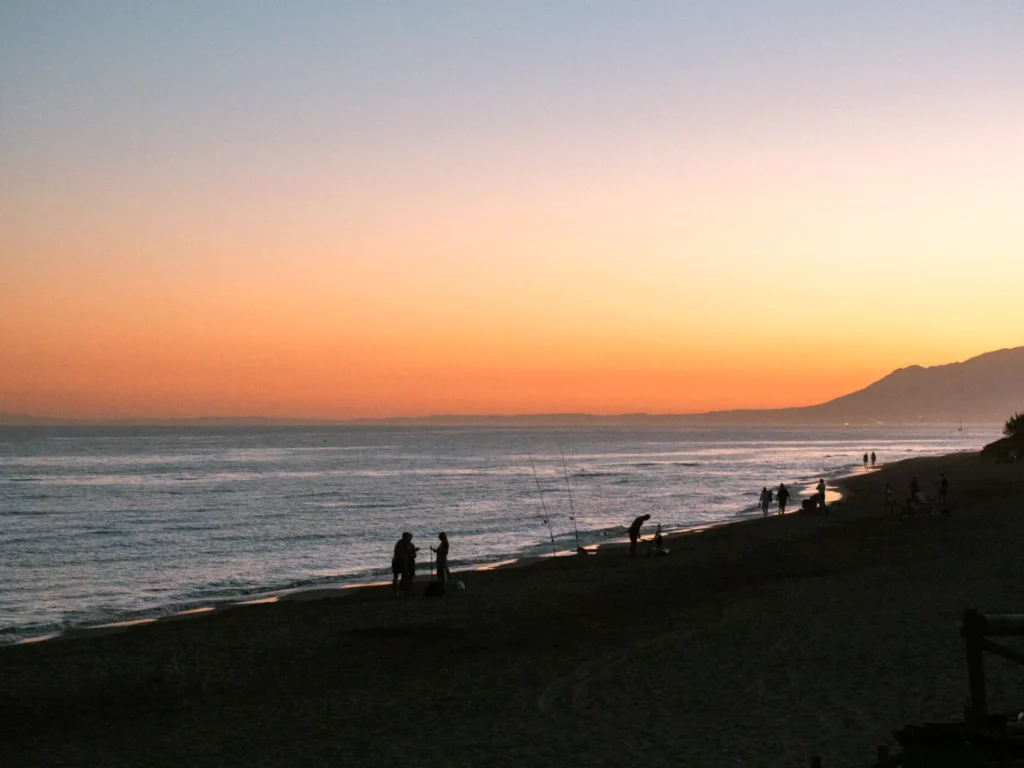 Sunset over the sandy beach at Cabopino, with people at the waterline and the coastline towards Marbella behind