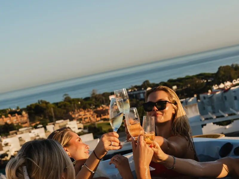 Three guests raising their glasses in the heated rooftop jacuzzi at sunset, with the Mediterranean behind
