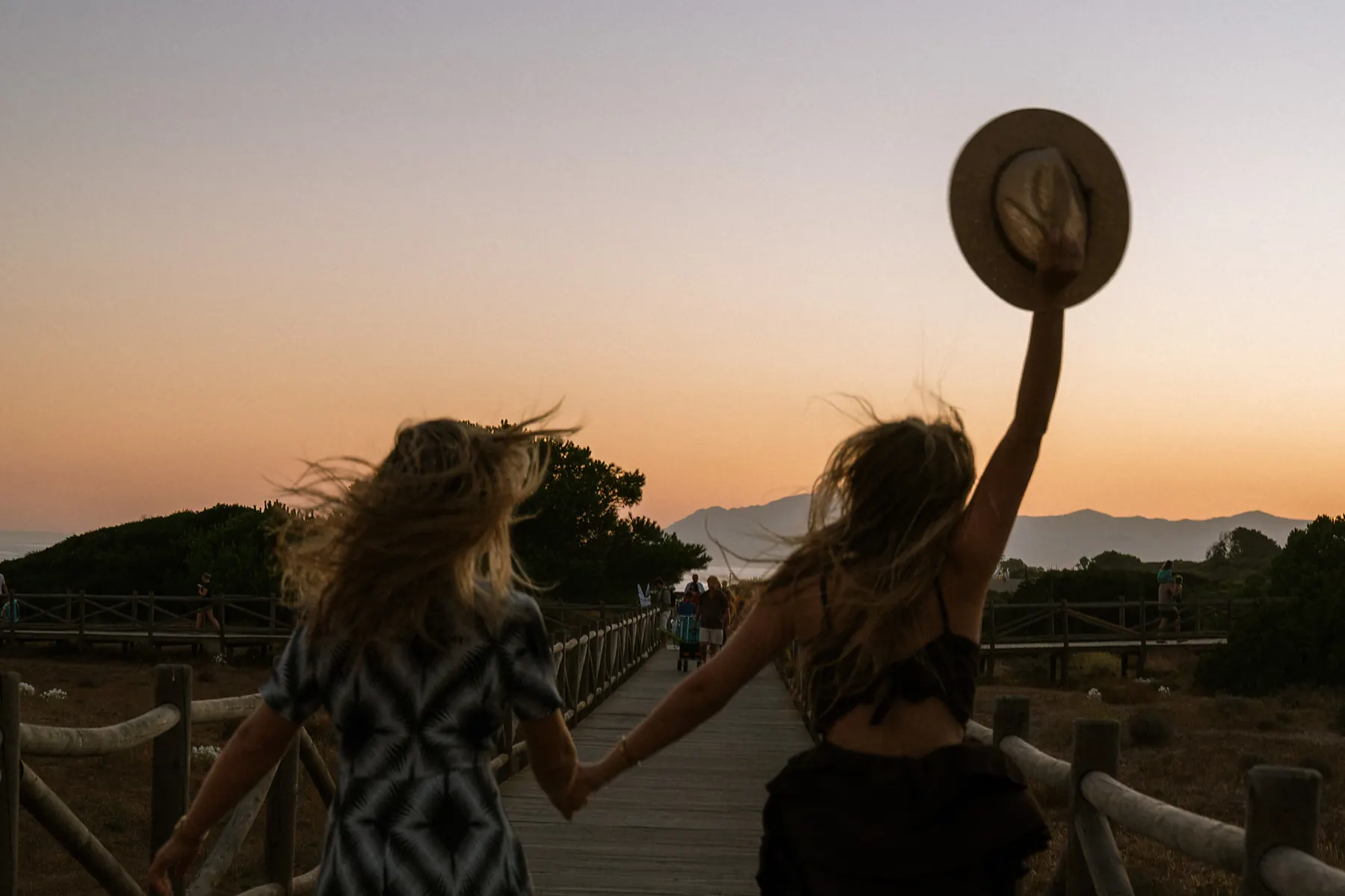 Two guests walking the boardwalk through the dunes at golden hour, one holding a straw hat in the air